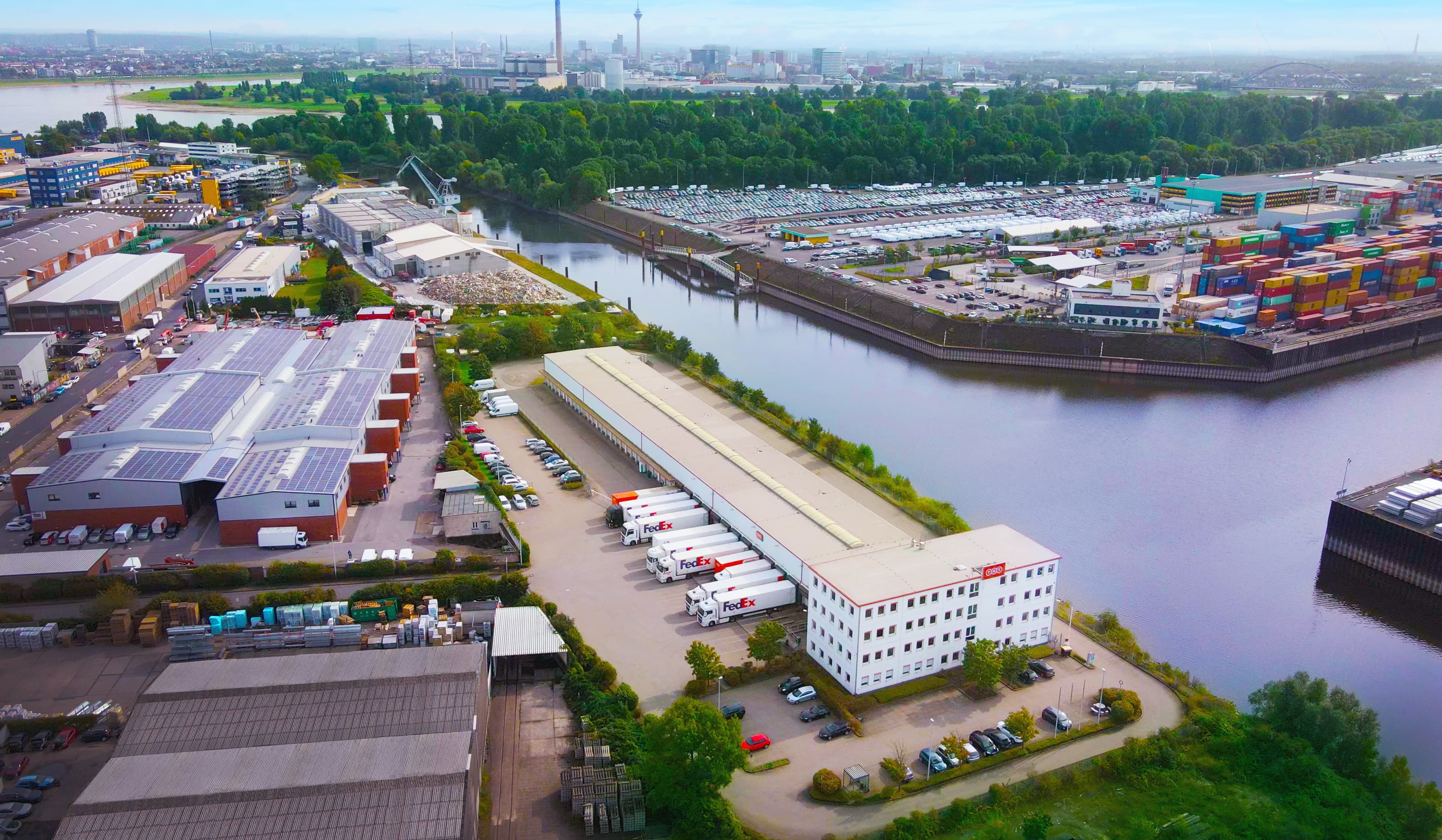 Aerial view of an industrial area with warehouses, parking lots, and a river. Numerous shipping containers and trucks are visible. City skyline in the background under a clear sky.
